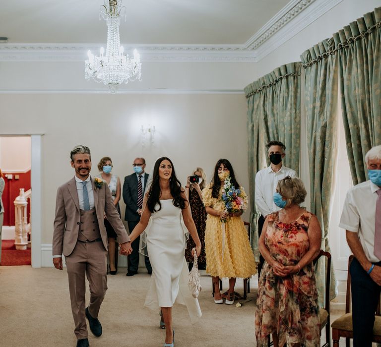 Bride in white strapless Rebecca Vallance Dress, veil and blue slingback heels carries ivory bridal bag whilst holding hands with groom in brown three piece Moss Bros suit and blue tie as they walk down the aisle at Harrogate wedding