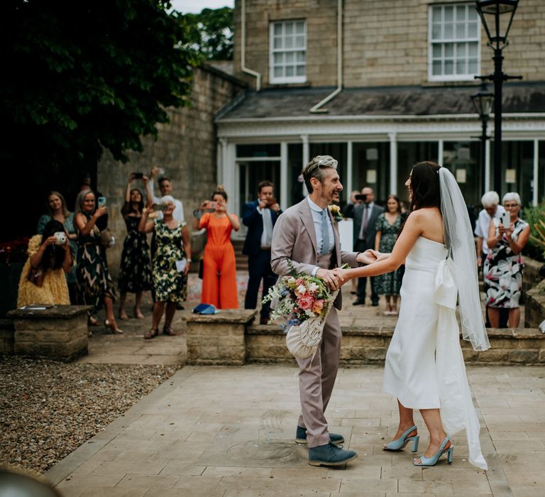 Bride in white strapless Rebecca Vallance Dress with bow detail at the back, veil and blue slingback heels holds multicoloured bouquet whilst dancing with groom in brown Moss Bros suit and blue tie in the courtyard at Hotel du Vin Harrogate after Harrogate wedding 