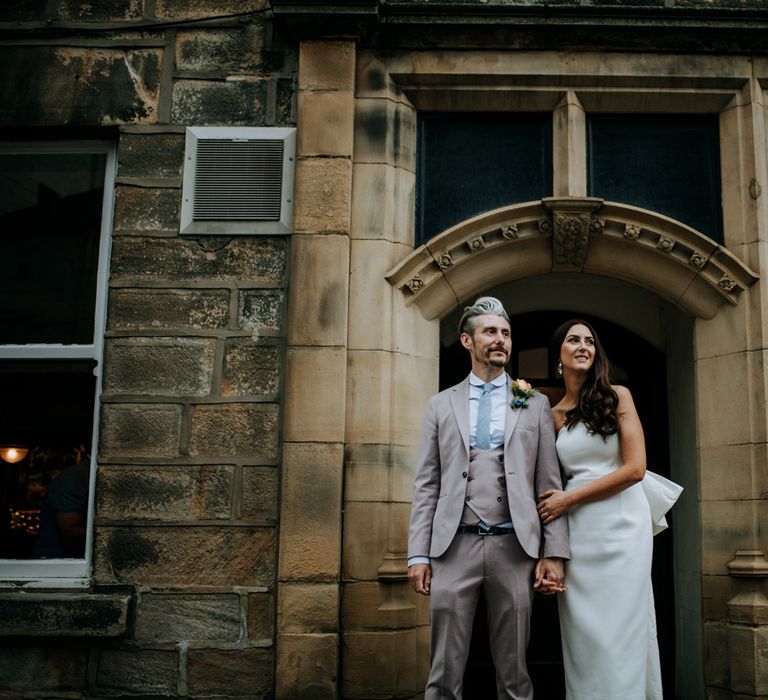 Bride in white strapless Rebecca Vallance Dress stands arm in arm with groom in light brown Moss Bros suit and blue tie on the stress of Harrogate after wedding