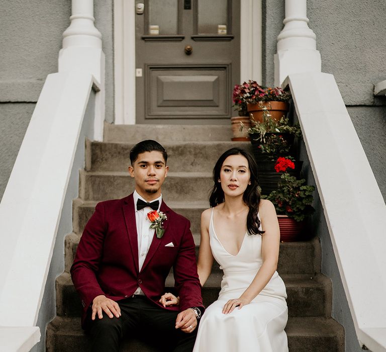 An Asian couple sit on steps for a wedding portrait. The bride wears a white dress and the groom wears a burgundy red jacket.