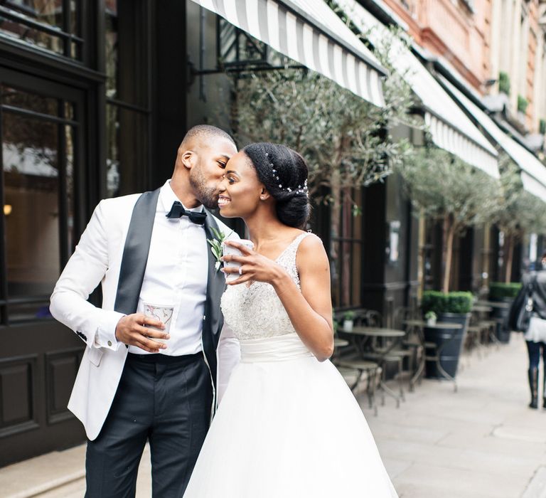 A Black couple walk down the street for wedding portraits. He wears a white tux jacket and she wears a long white dress. They hold takeaway coffee cups.