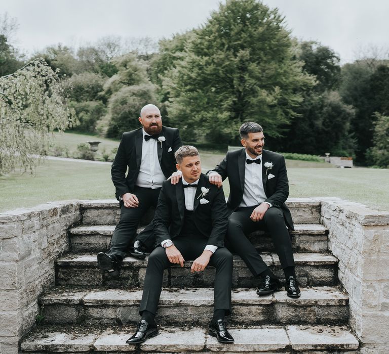 Groom and two groomsmen in black tuxedos sit on stone steps in the grounds of Came House Dorset as they look off camera