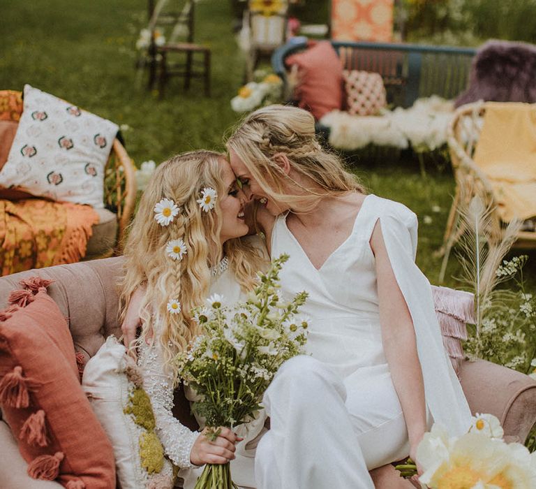 Bride in a jumpsuit with half up half down hair sitting on her brides lap on a retro sofa at outdoor wedding ceremony 