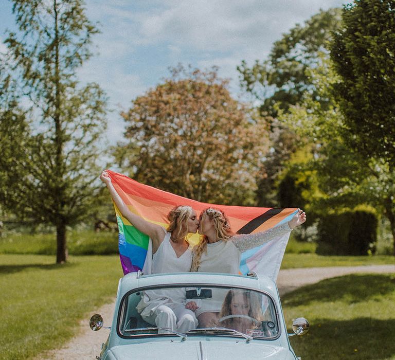 Two brides kissing and holding a Pride flag on the back of the light blue fiat 500 car 