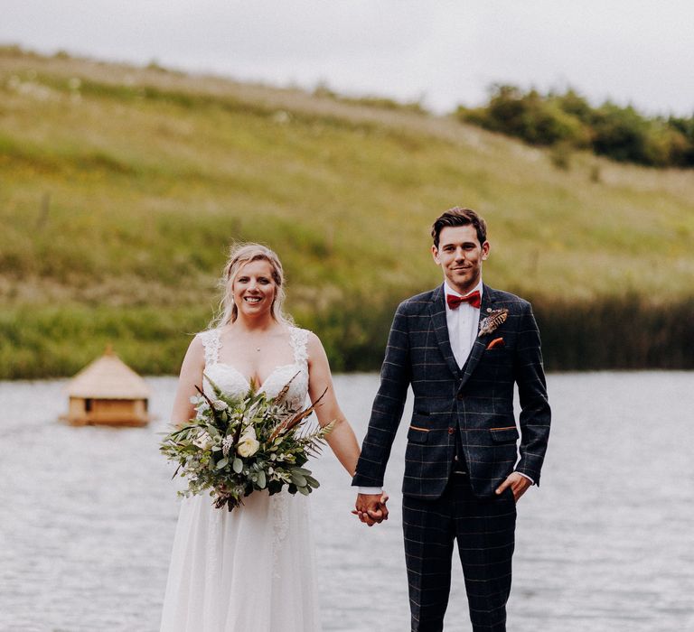 Bride & groom hold hands as they stand together in front of the lake, as bride holds floral bouquet complete with feathers 