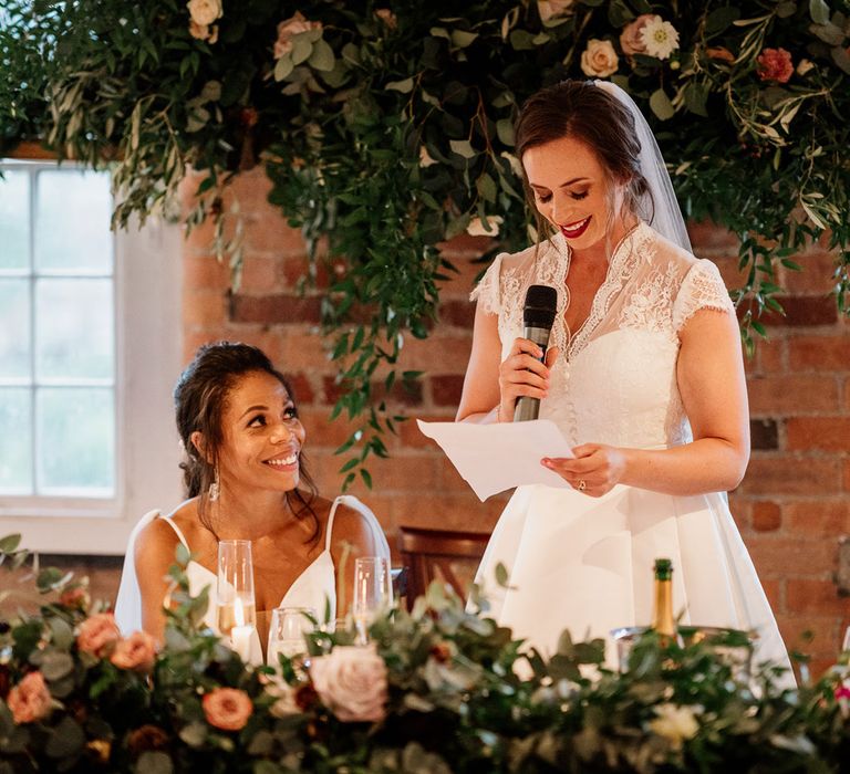 Bride sat down in white cami dress with shoulder veil details looks up at bride in white lace top capped sleeve wedding dress and veil as she holds microphone and makes speech during wedding reception at The West Mill Derby