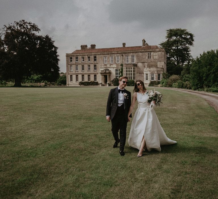 Bride & groom walk through the grounds of Elmore Court whilst wearing sunglasses on their wedding day