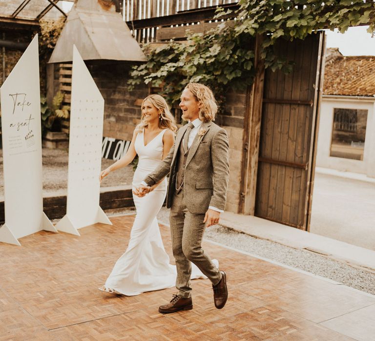 Bride in white Made With Love wedding dress holds hands with groom in sage green suit as they walk into glasshouse wedding breakfast at Anran House