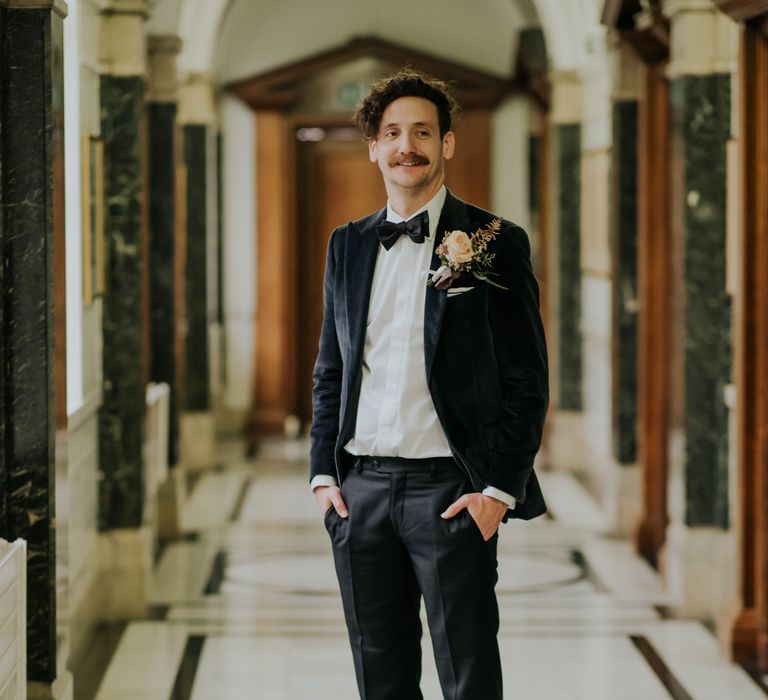 Groom looks dapper in deep velvet suit complete with bow tie and floral buttonhole 