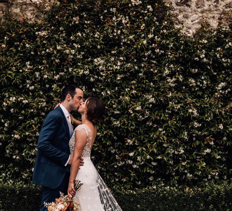 Bride in white open back Rime Arodaky wedding dress with lace train holding dried bridal bouquet kisses groom in blue suit outside at summer wedding in Dorset