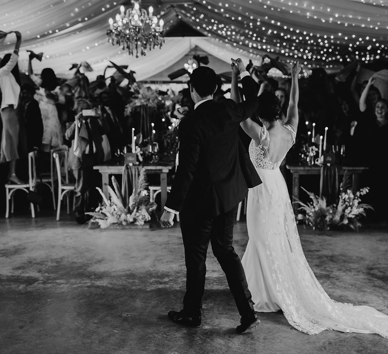 Bride in open back Rime Arodaky wedding dress and groom in blue suit enter wedding breakfast as guests stand on chairs and wave napkins during summer wedding in Dorset