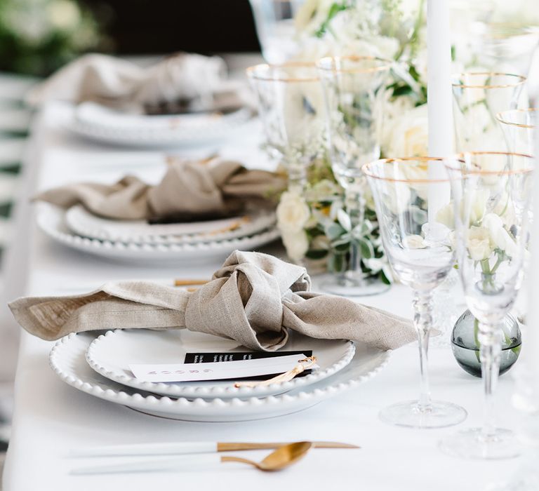 Three table settings at a black and white wedding .Each setting has a neutral coloured tied napkin, gold cutlery, place tag with gold tassel and tall white tapered candles.