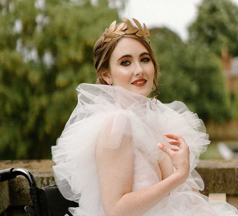 Bride in a tulle wedding dress and gold crown sitting on the balcony at Bourton Hall in her wheelchair with Crocheted wheelchair covers