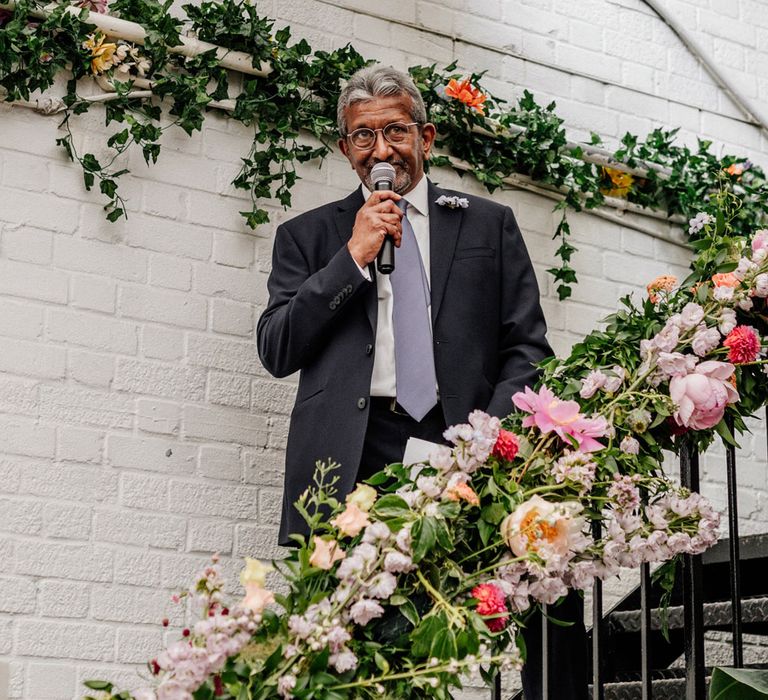 Father of the bride in dark suit and light blue tie stands on metal stairs with floral banister decoration making speech at Loft Studios London