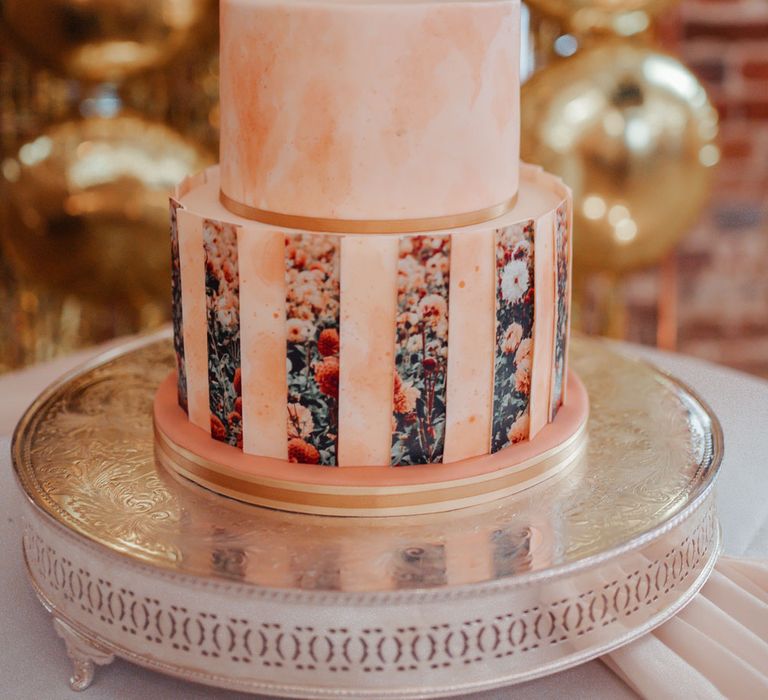 Blush pink marble effect two tier wedding cake with floral photo detailing and orange flower on top sat on gold platter in front of golden balloons at Wasing Park wedding reception