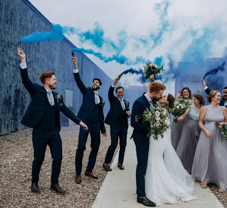 Bride and groom kissing surrounded by their wedding party in grey bridesmaid dresses and navy suits holding blue smoke bombs 