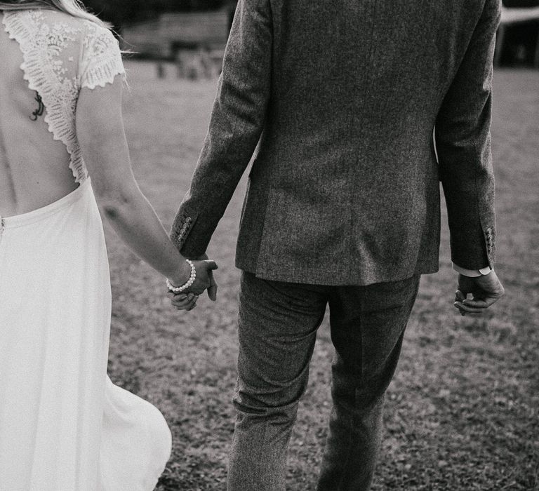 Bride in lace top open back wedding dress wearing pearl bracelet holds hands with groom in grey herringbone suit as they walk outside during late summer wedding at Wellington Wood Norfolk