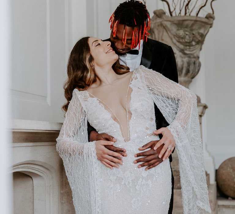 Black groom in a tuxedo embracing his bride in an embellished lace wedding dress with plunging neckline and long bell sleeves