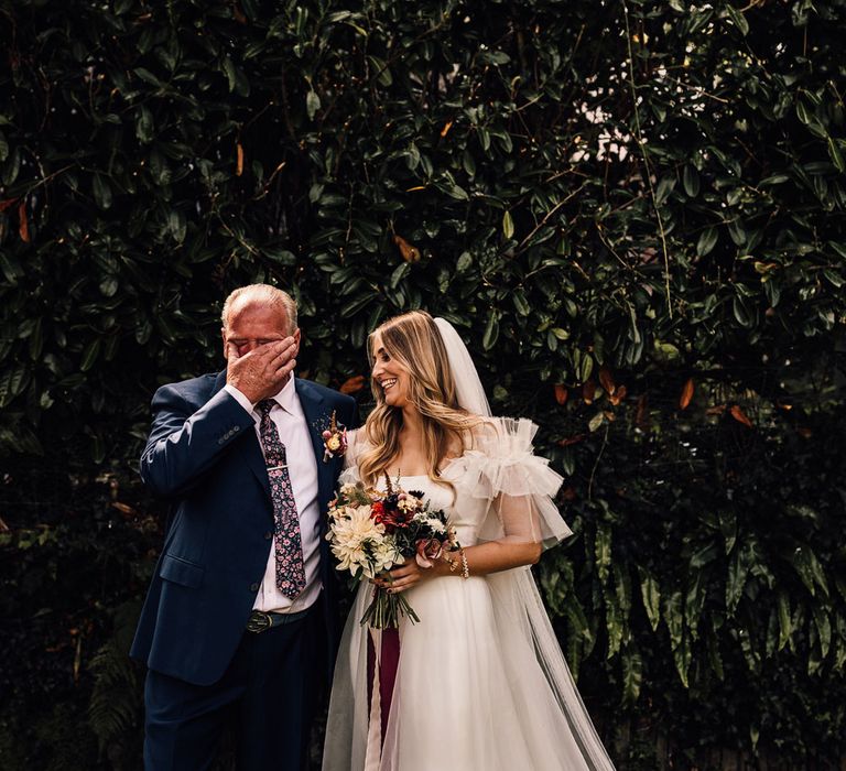 Bride in white Halfpenny London Mayfair dress stands outside holding mixed bridal bouquet with burgundy ribbon with emotional father in blue suit before church wedding ceremony in Cornwall