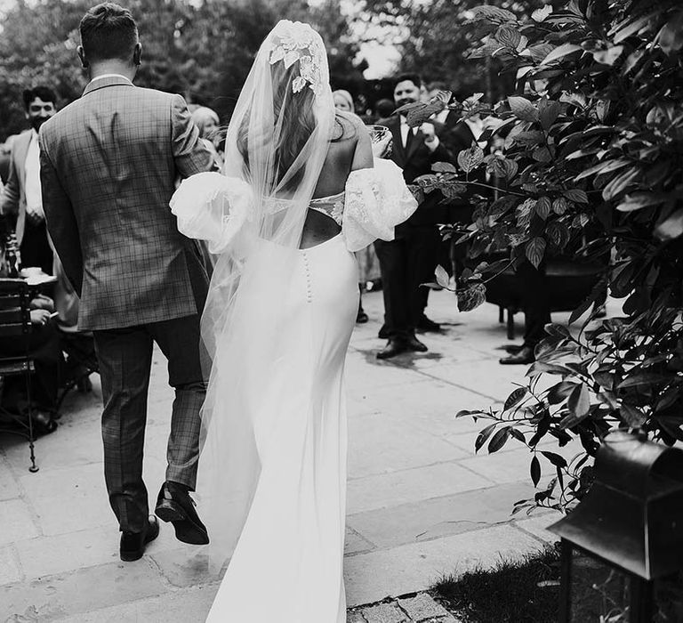 Bride & groom walk together in black & white image as bride wears lace embellished veil 