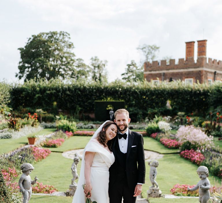 Bride & groom stand with one another in lush gardens in front of Hampton Court Palace