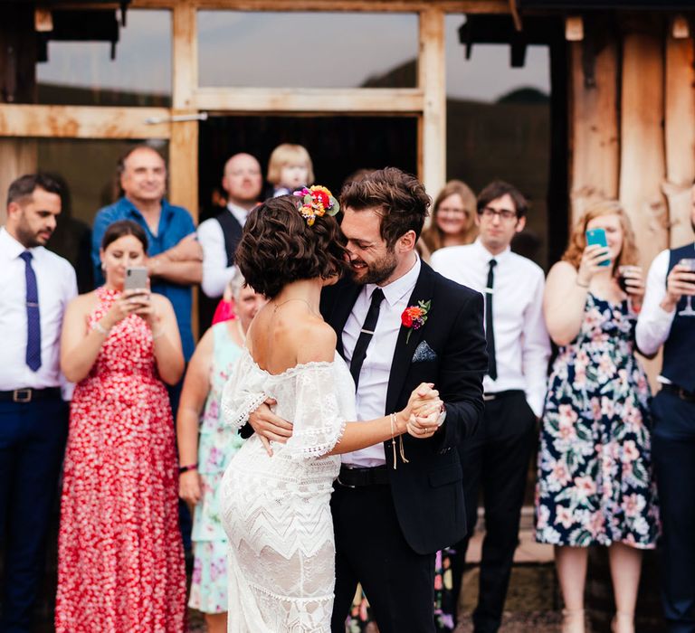 Bride in off the shoulder Grace Loves Lace wedding dress, dusty pink block heels and colourful flower crown dances with groom in black suit and colourful floral buttonhole outside The Bridal Barn wedding venue as wedding guests watch