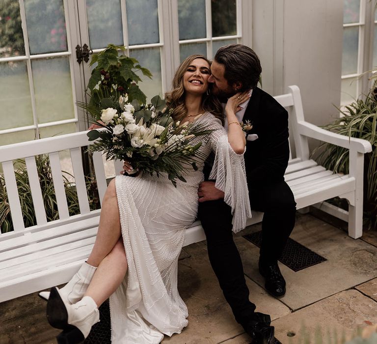 Bride in beaded Eliza Jane Howell wedding dress with white boots sitting on a bench with her groom in a tuxedo at Tatton Park 