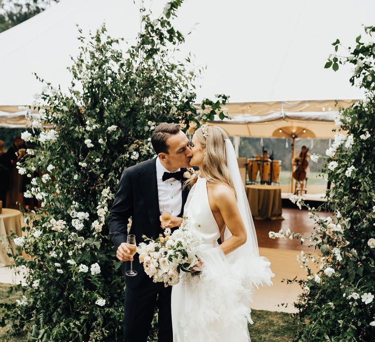 Bride in halterneck Halfpenny London wedding dress and ruffle veil holding white and green bridal bouquet kisses groom unblock tie as they stand outside of tent wedding reception 