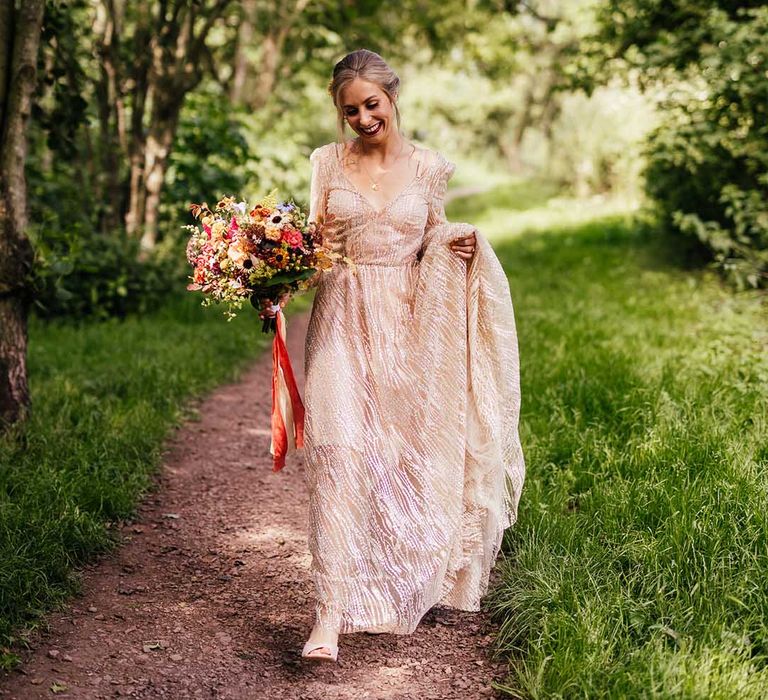 Blonde bride holds her sparkling bridal gown whilst holding colourful bouquet tied with red ribbon