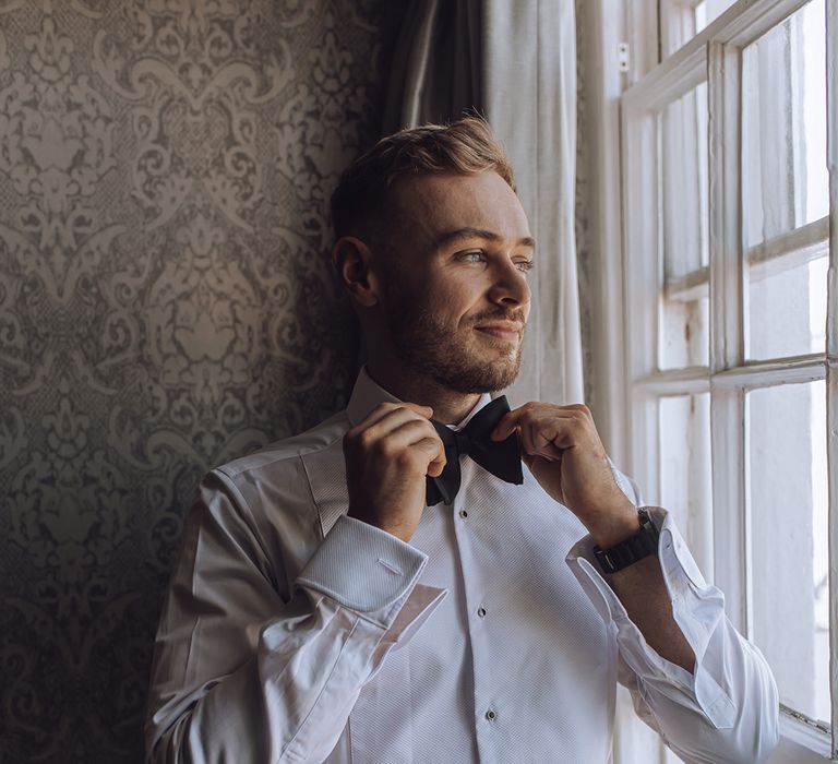 Groom does up his black tie as he looks out the window on his wedding day