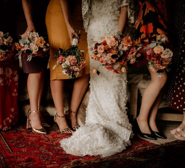 Bride in wedding dress with applique flowers stands with bridesmaids in mismatched bridesmaids dresses all holding mixed pink, orange and white bridal bouquets