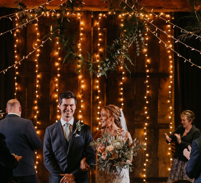 Bride in lace and tulle wedding dress holding large mixed bridal bouquet walks down the aisle with groom in dark suit and light blue tie for fairy-lit wedding ceremony