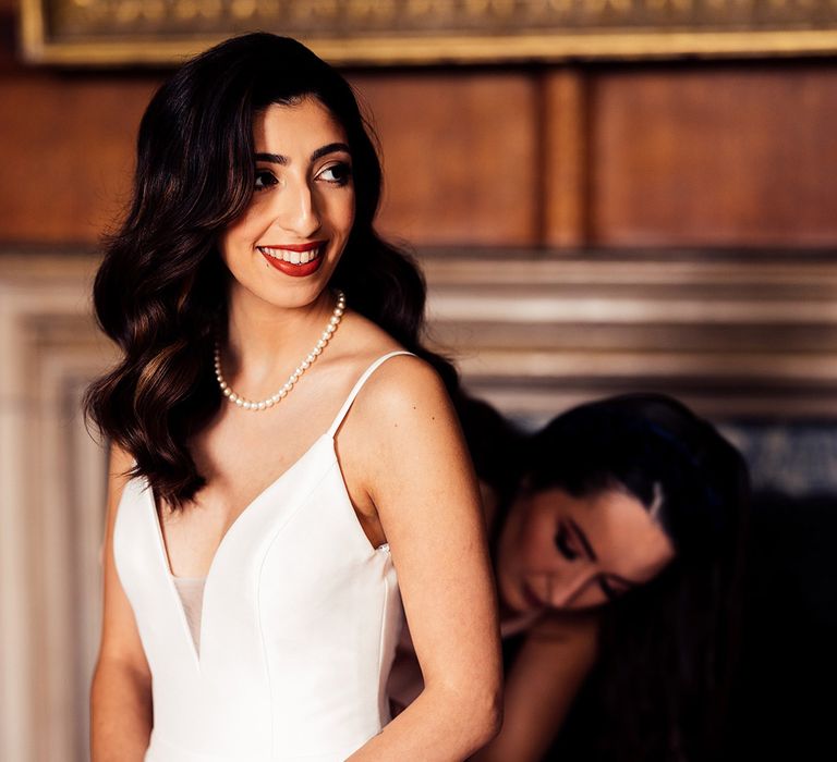 Bridesmaid in deep v wedding dress with pearl necklace stands in bedroom before wedding as bridesmaid adjusts the back fastenings