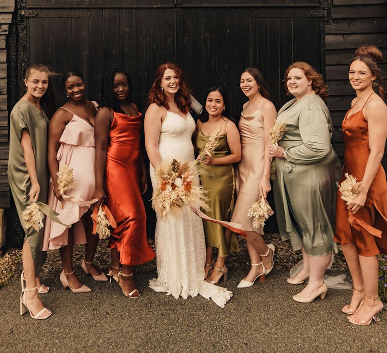 Bride stands with her bridesmaids who wear Autumnal coloured bridesmaids gowns in different colours and styles 