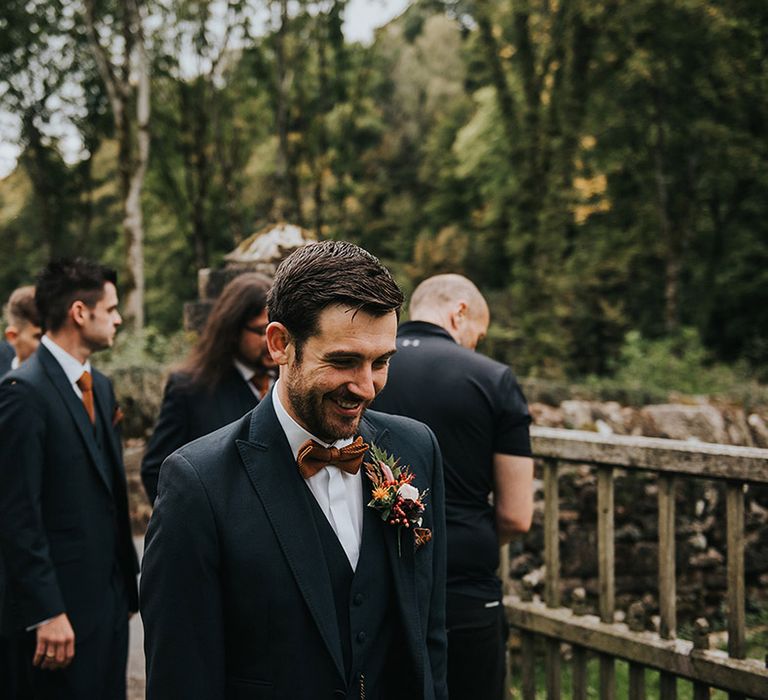 Groom in a navy blue three-piece wedding suit with autumn buttonhole flowers and brown bow tie 