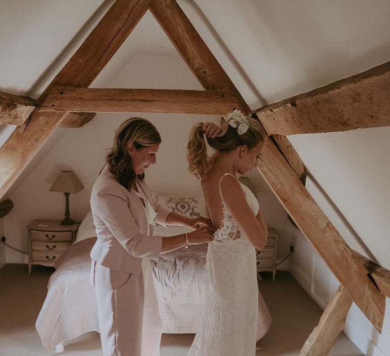 Mother of the bride in muted pink power suit does up the bride's lace wedding dress on the morning of the wedding