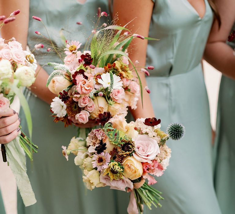 Bridesmaids in v-neck sage green satin dresses holding wildflower bouquets with pink, red, white and orange flowers