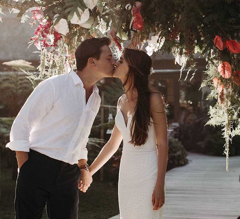 Groom in white shirt and black trousers kisses bride in fitted v-neck wedding dress next to tropical flowers