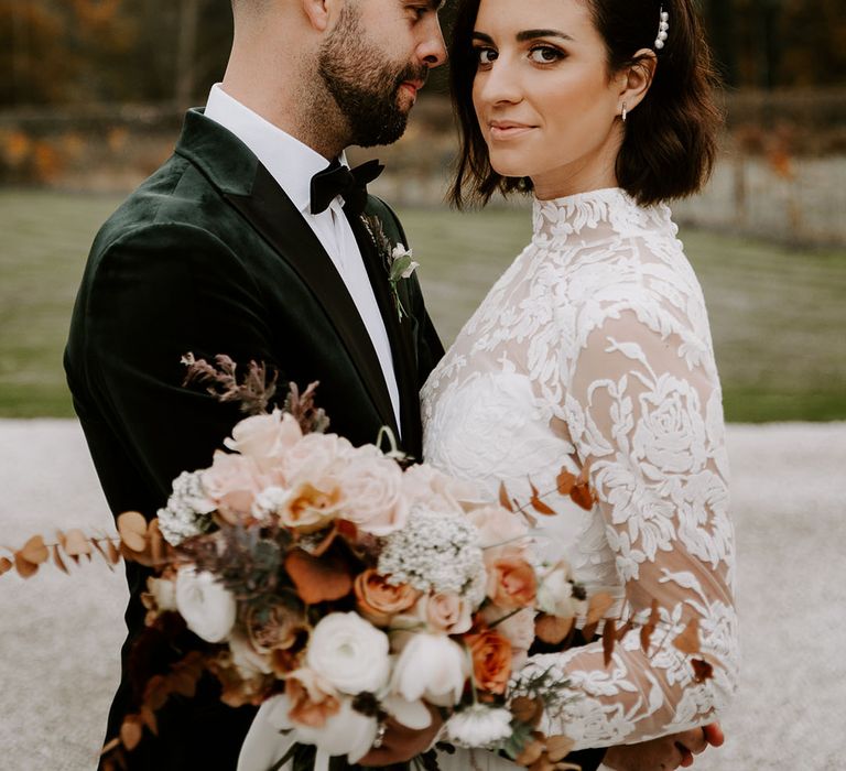 Bride in high neck long sleeve lace wedding dress and peal hair accessory hold autumnal wedding bouquet as she holds onto groom in velvet black tie