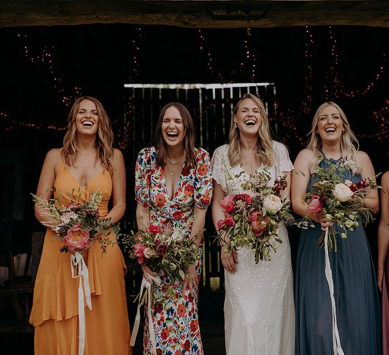 Bride and bridesmaids laugh together with pink, red and white bouquets and bridesmaids all in different colour and style dresses