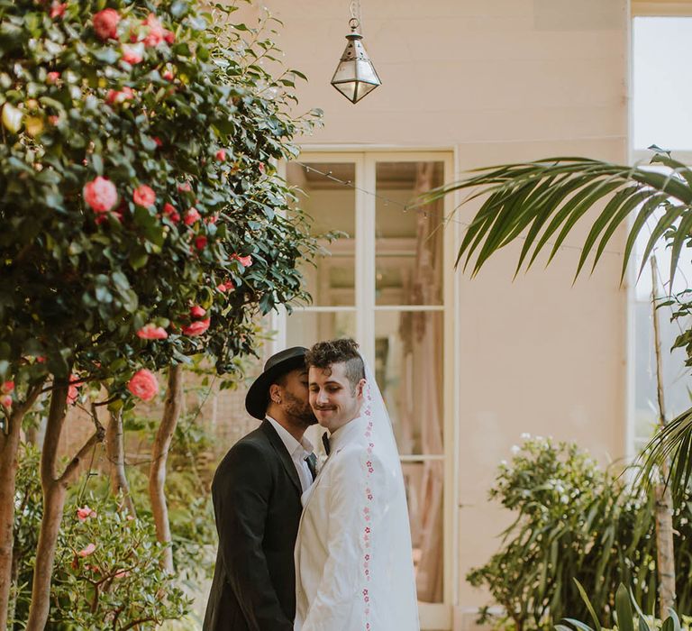 Groom in a black suit, fedora hat and boots kissing his partner in a white groom suit and patterned edge veil at Prestwold Hall Barns