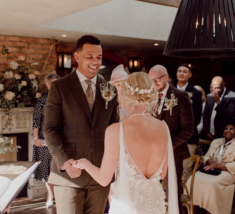 Groom in brown checkered suit and patterned tie smiles down at bride in lace open back wedding dress with braided updo