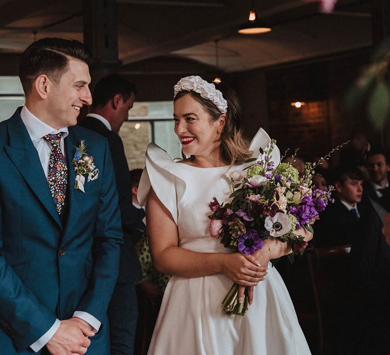 Groom in blue suit and floral tie smiles at the bride holding her purple wedding bouquet in a Jesus Peiro wedding dress