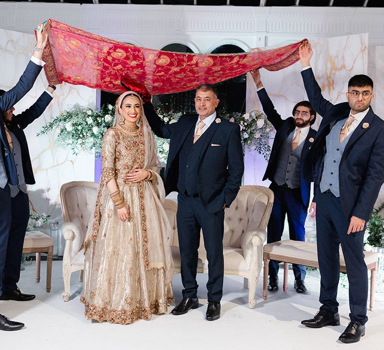 Bride & groom stand beneath brightly coloured fabric held up by wedding guests after Nikkah & Walima