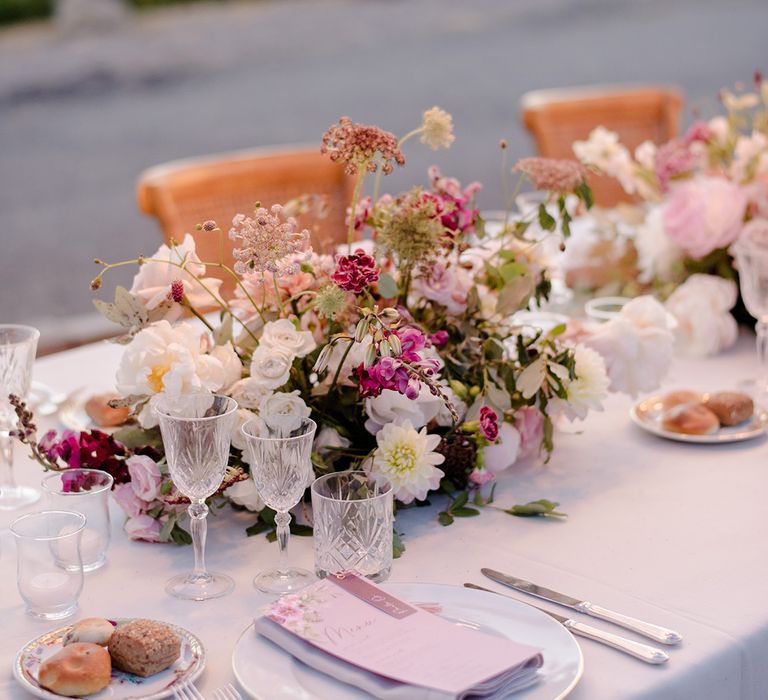 Lavender and pink florals sit in bouquet on table beside lavender stationery and crystal glass for outdoor reception in Tuscany 