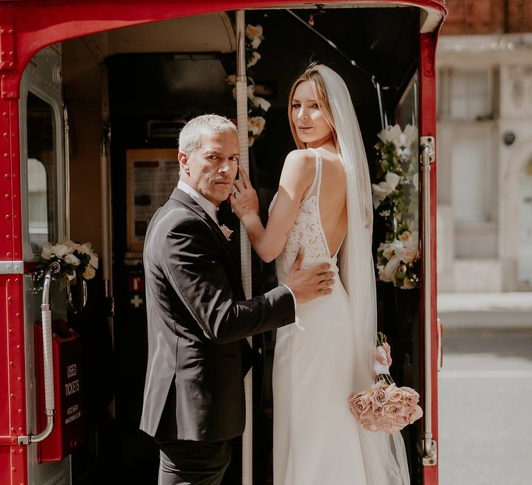 Groom in black tie rests his hand on the bride's back who wears a beaded wedding dress with a pink rose round bouquet 