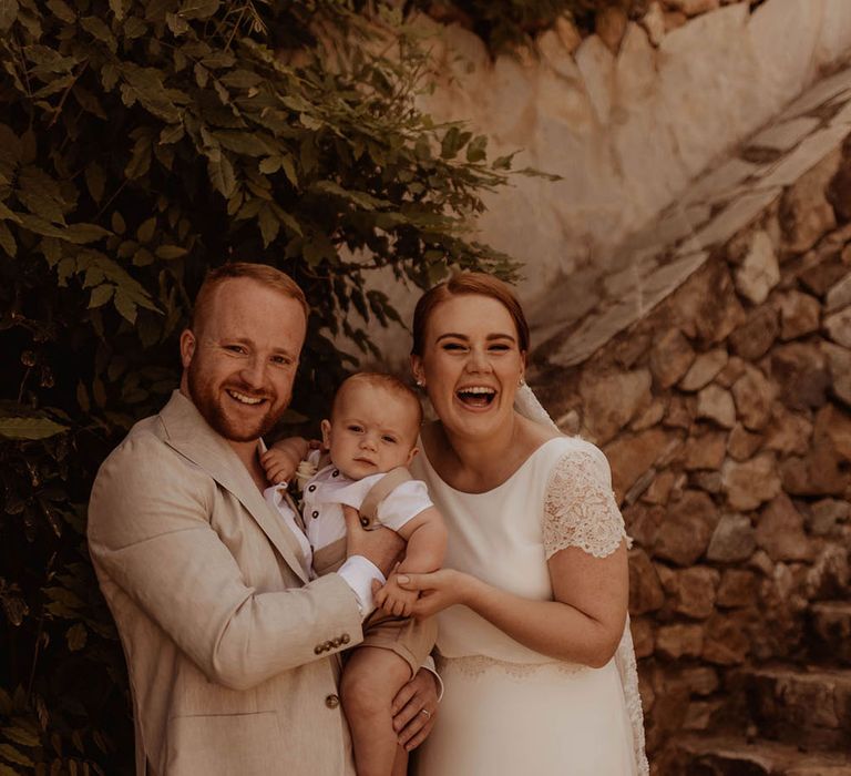 Bride & groom hold their baby as bride wears gown complete with lace sleeves and waistband 