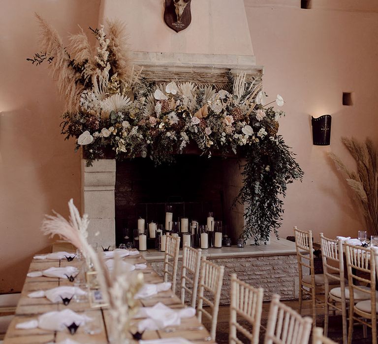 Rustic reception with wooden banquet tables in front of white fireplace surrounded by florals and pillar candles