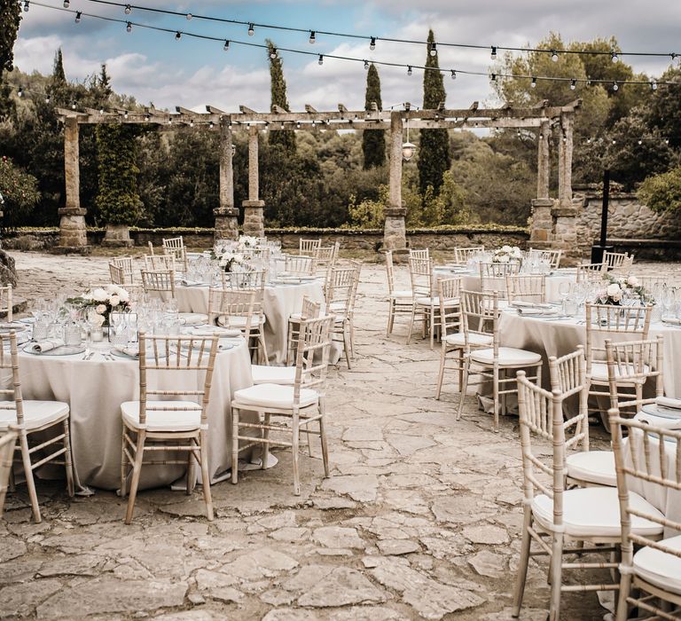 Chic and neutral outdoor wedding reception complete with round tables and white linen tablecloths, finished with small pastel bouquets and white wicker chairs 