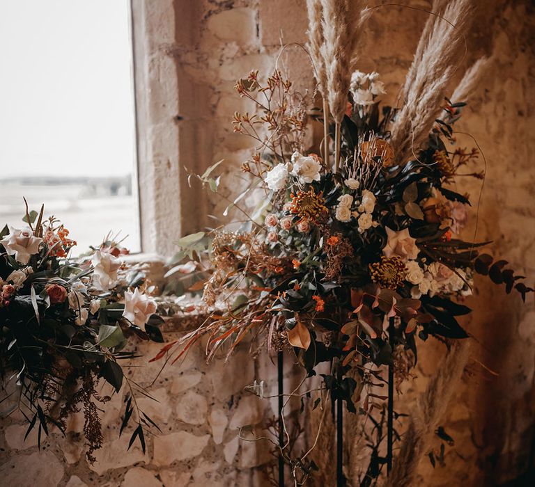 Large dried floral arrangements with pops of orange, white and pale pinks alongside pampas grass surrounded by white pillar candles